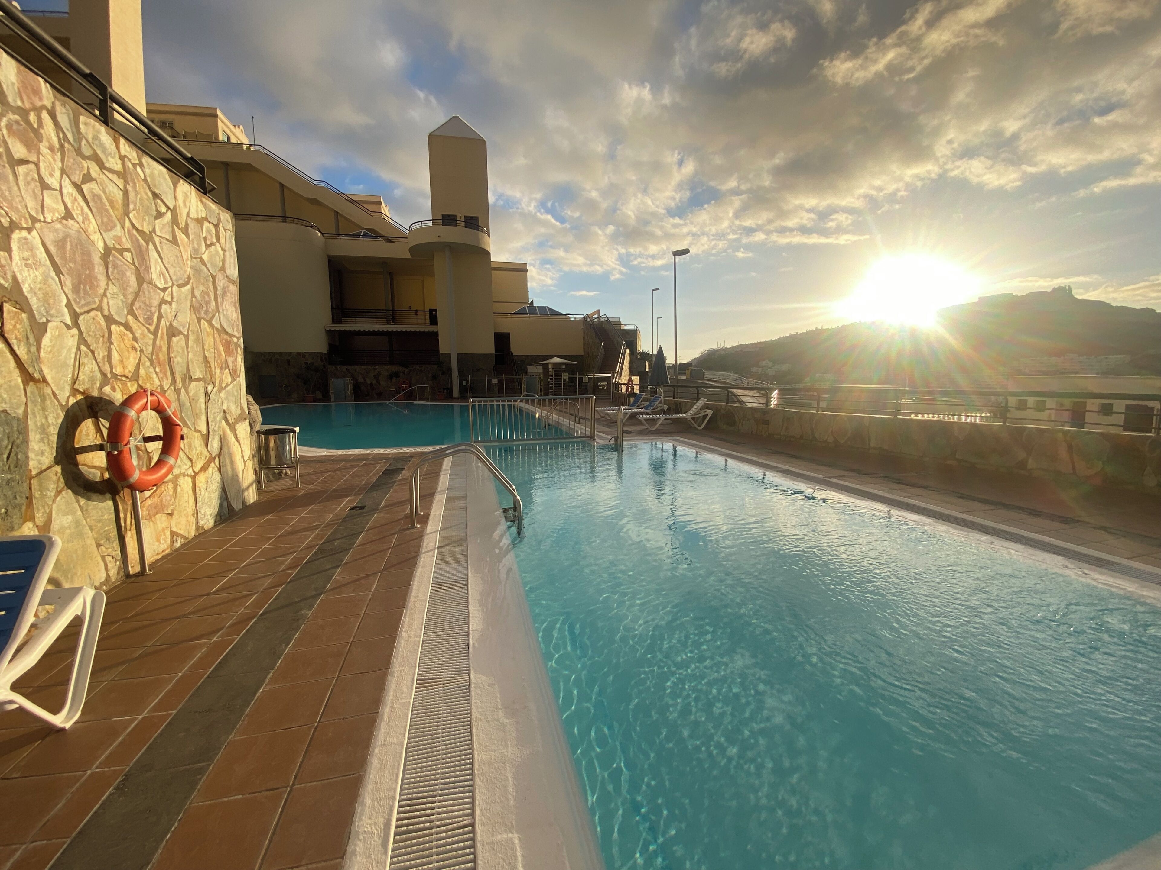 A swimming pool with a stone wall and lifebuoy, surrounded by a building with a balcony.