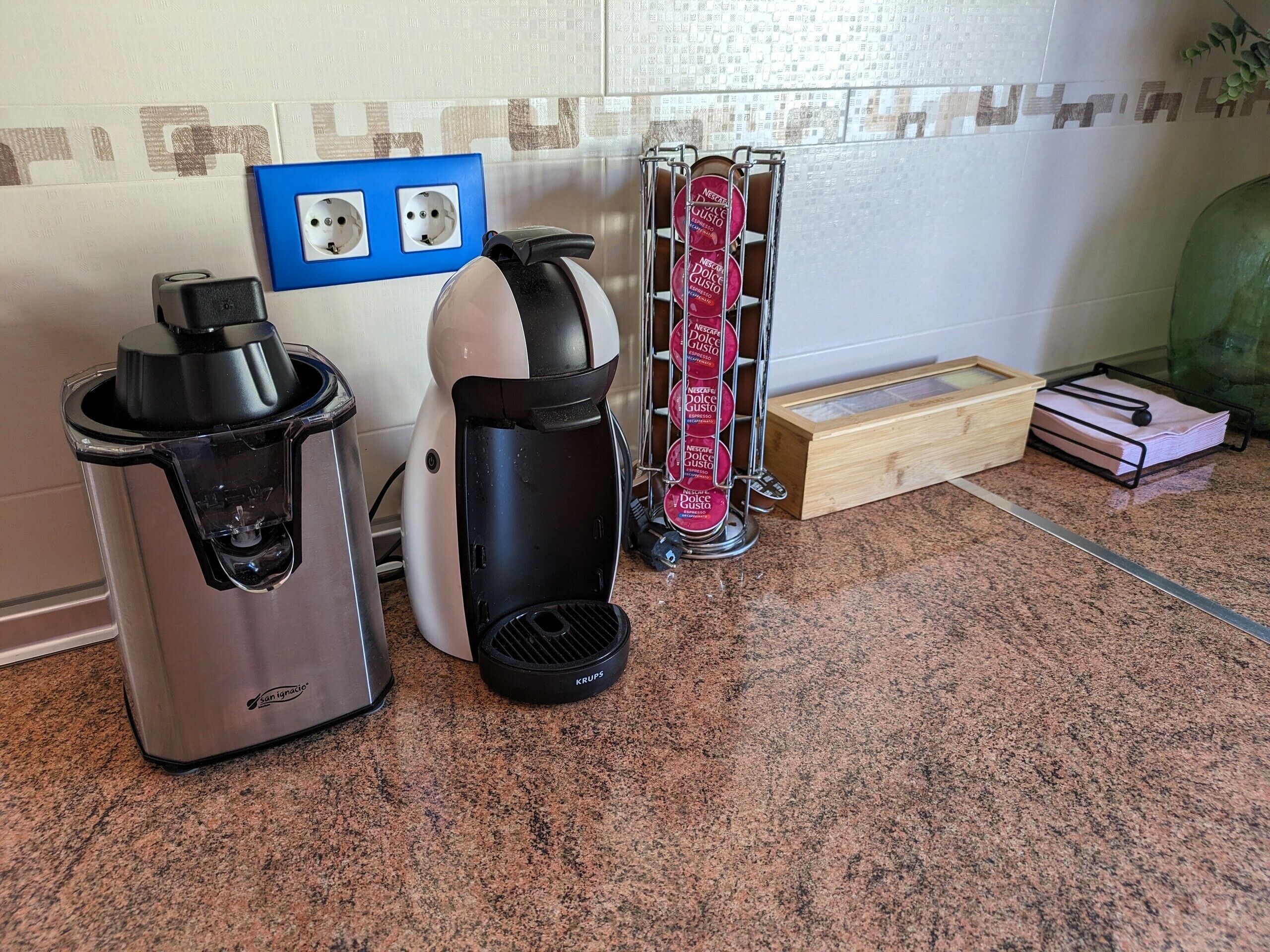 Een keuken met een koffiezetapparaat, een waterkoker en een koffieautomaat. Er is een blauw stopcontact en een houten doos op het aanrecht.