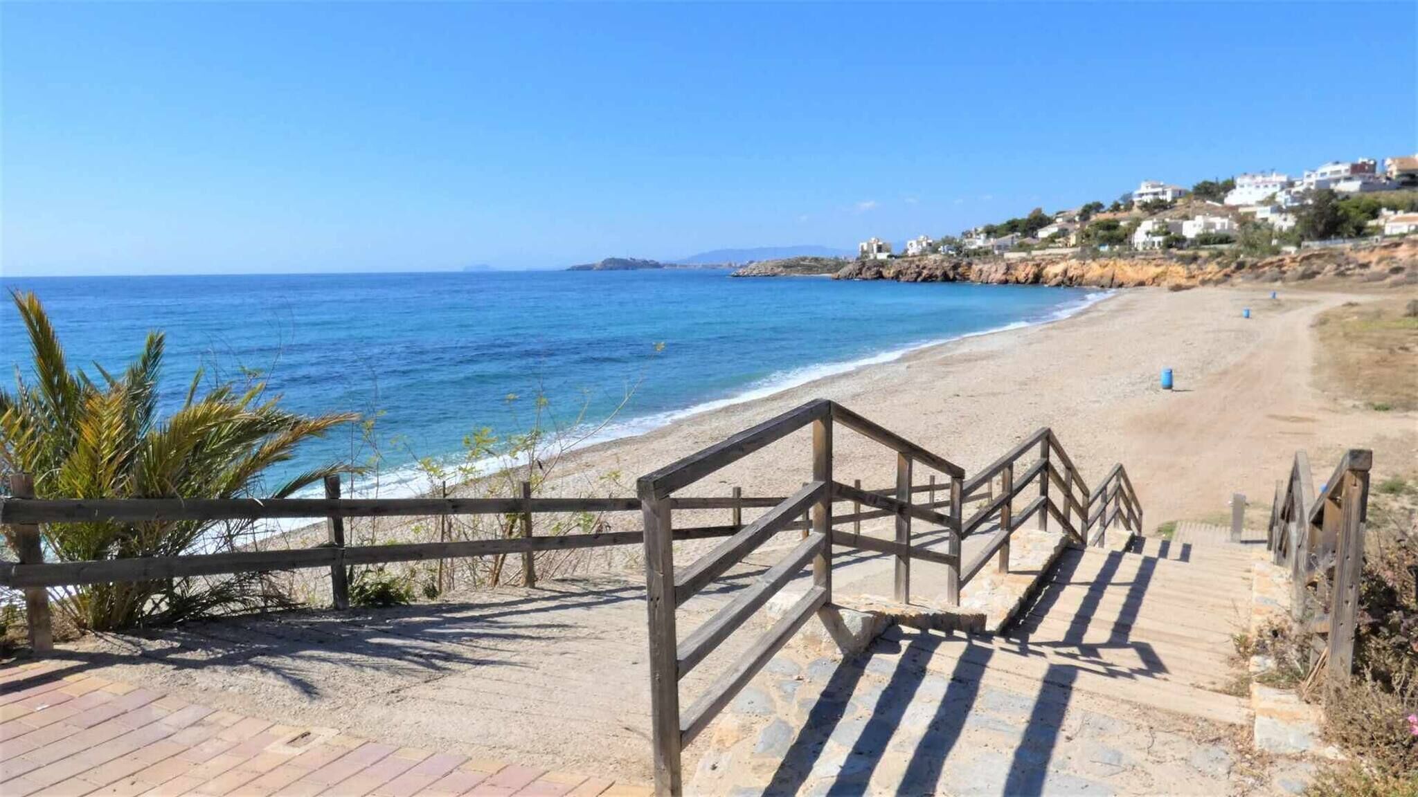 Una platja amb baranes de fusta que condueixen a la riba, un cel blau i clar, i edificis en un turó al fons.