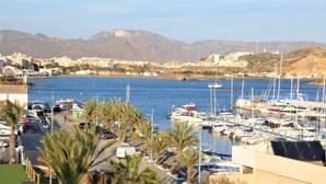 Uma marina com barcos atracados, vista clara do mar e montanhas ao fundo.