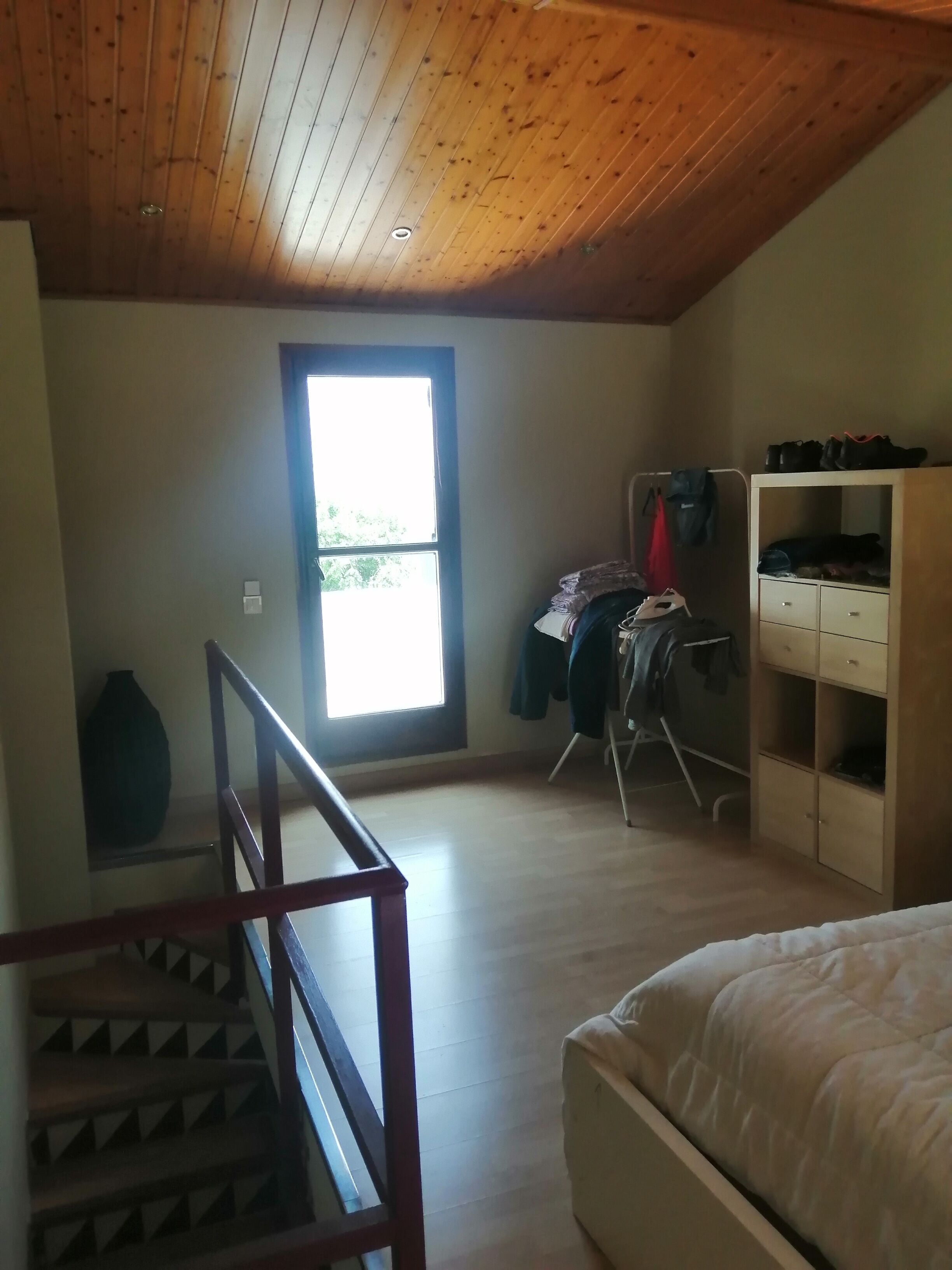 A bedroom with a wooden ceiling, a bed, a wardrobe, and a desk with clothes hanging.