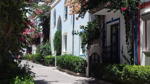 A narrow street with white buildings, arched doorways, and flowering plants.