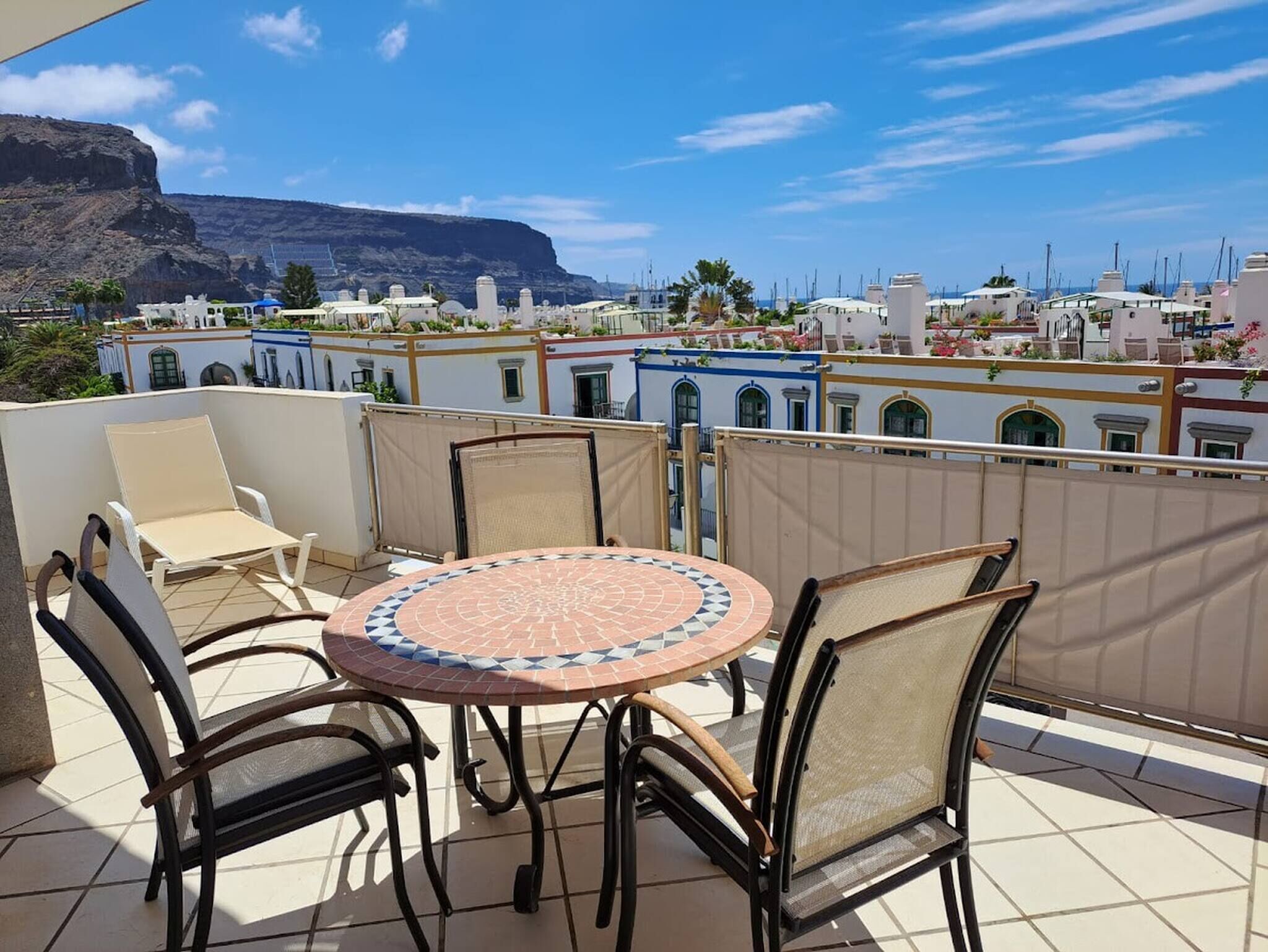 Une terrasse avec une table ronde et quatre chaises, d’où l’on a une vue sur une ville c ôtière, avec des bâtiments et une montagne en arrière-plan.