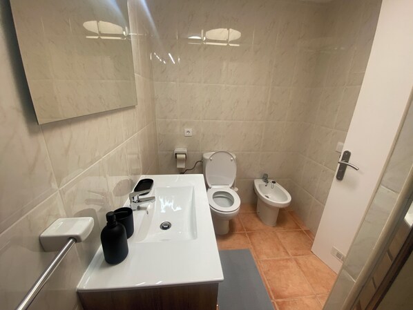 Un baño moderno con un lavabo blanco, un inodoro y un bidé. Hay un espejo sobre el lavabo y un perchero a la izquierda.