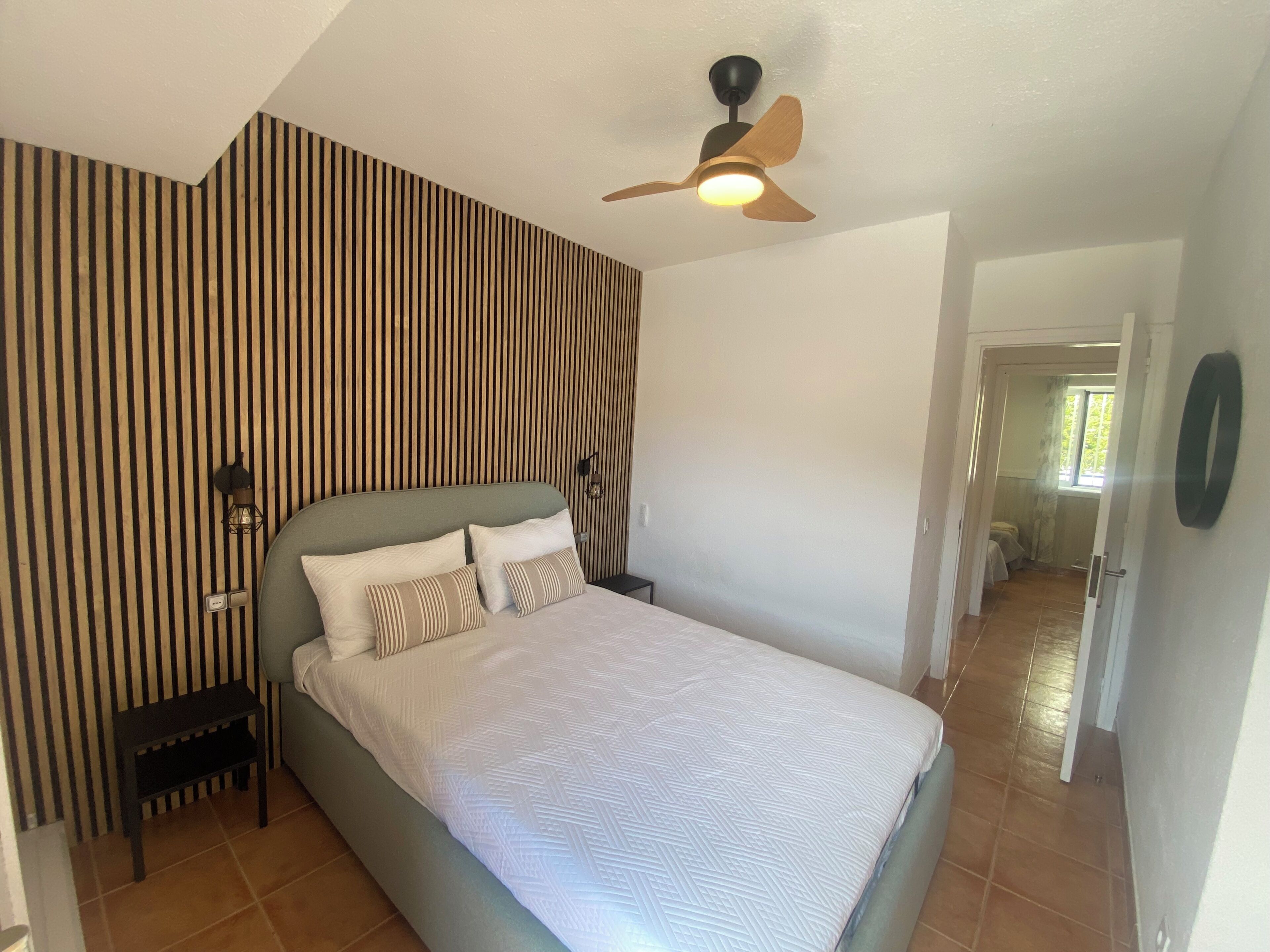 A bedroom with a bed, a nightstand, a wall-mounted light, a ceiling fan, and a door leading to another room.