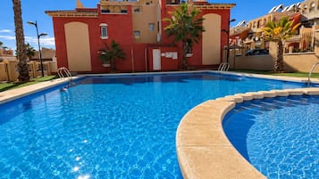 Une piscine à bordures courbes, entourée d’un bâtiment aux murs rouges et beiges, de palmiers et sous un ciel d’un bleu limpide.