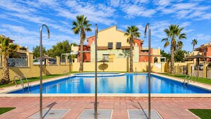 A swimming pool with a clear blue sky, surrounded by palm trees and a building with a red roof. - Beach & Pool Av Mancha (Torrevieja)