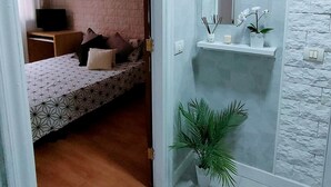 Une chambre à coucher avec un lit, des oreillers, une télévision et un petit bureau. Il y a une salle de bain avec un miroir, une étagère et des plantes.