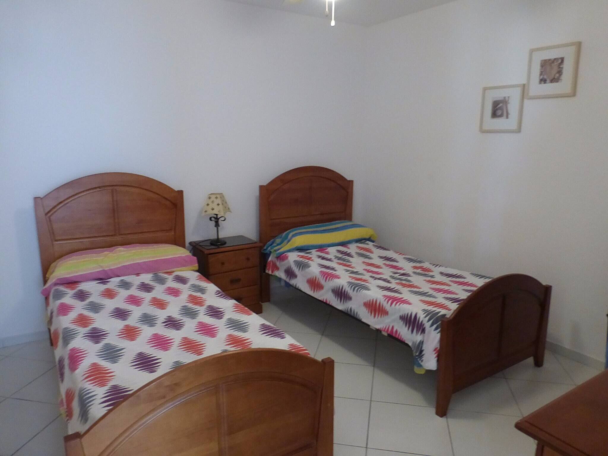 Una habitación pequeña y limpia con dos camas de madera, una mesita de noche con lámpara y una pequeña mesa.
