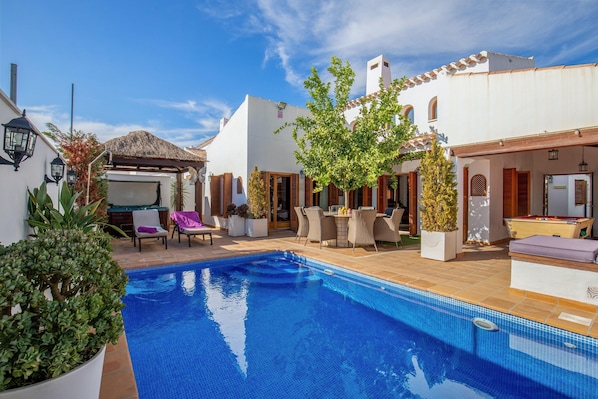 A swimming pool with lounge chairs and a table, surrounded by white buildings with wooden shutters and a thatched roof structure. - Special-offer Luxuriousvilla, Baños y Mendigo, Spain-perfect for Families&groups (Murcia)