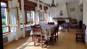 A dining area with a table set for four, surrounded by wooden chairs. There is a fireplace with a stone mantle, and a chandelier hangs from the ceiling.