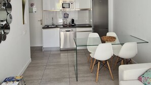 A modern kitchen with white cabinets, a stainless steel refrigerator, and a microwave. There is a dining area with white chairs and a glass table. - Luminoso Bilocale Vicino Al Mare (La Oliva)