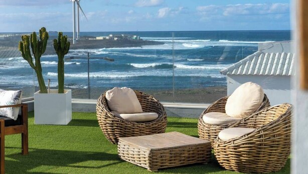 A terrace with wicker chairs and cushions, a cactus plant, and a view of the ocean and wind turbines. - Luminoso Bilocale Vicino Al Mare (La Oliva)