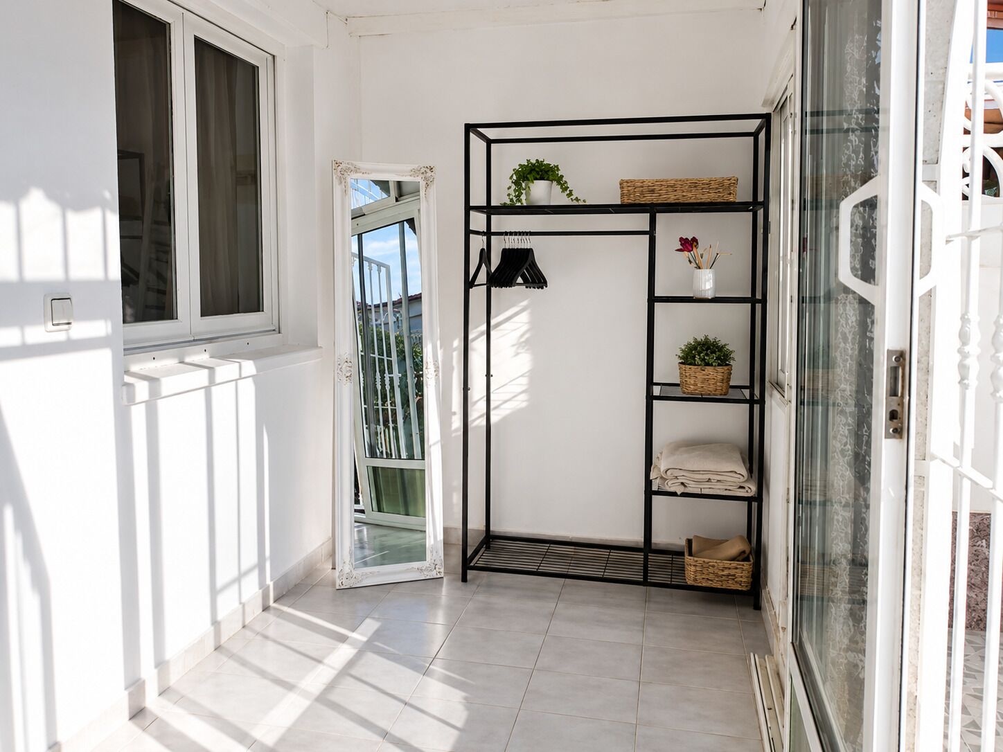 A hallway with a white door, a black metal shelf unit with baskets and a potted plant, and a window with a view of greenery.