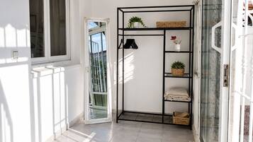A hallway with a white door, a black metal shelf unit with baskets and a potted plant, and a window with a view of greenery.