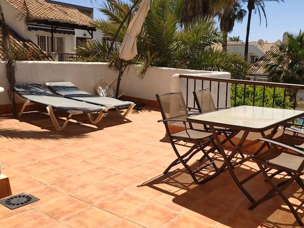 A terrace with a table and chairs, a sun lounger, and a view of palm trees and buildings.