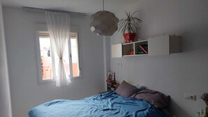 Een slaapkamer met een bed, een raam met gordijnen, een plank met boeken en een kamerplant.