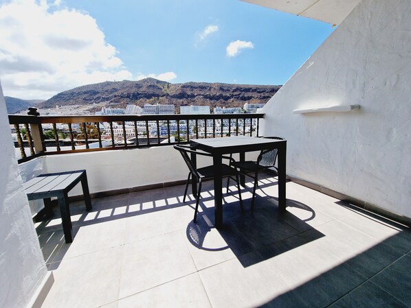 A balcony with a table and chairs, a view of a cityscape and mountains in the background.