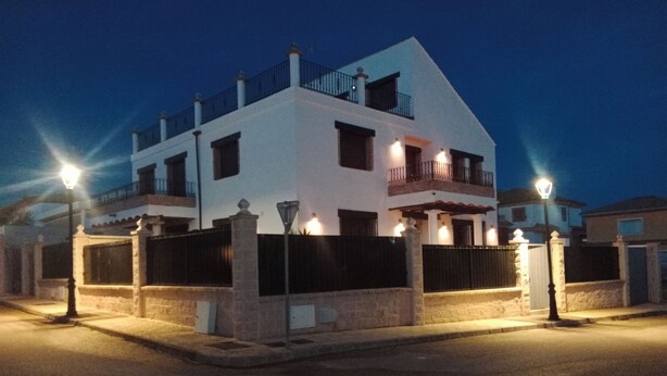 A modern two-story house with a balcony on the second floor, surrounded by a gated fence and street lamps.