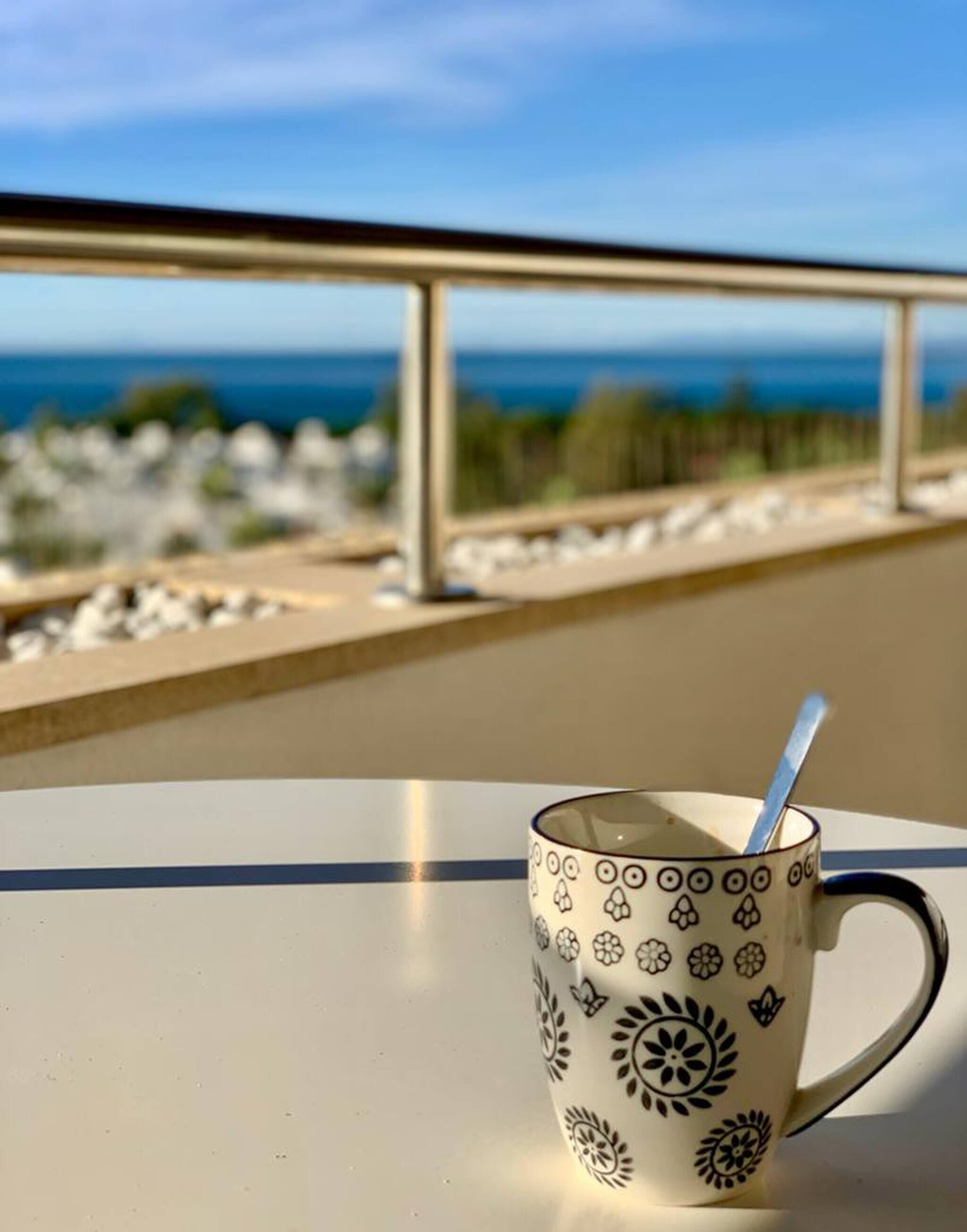 A cup with a floral pattern on a table with a view of the sea and a railing.