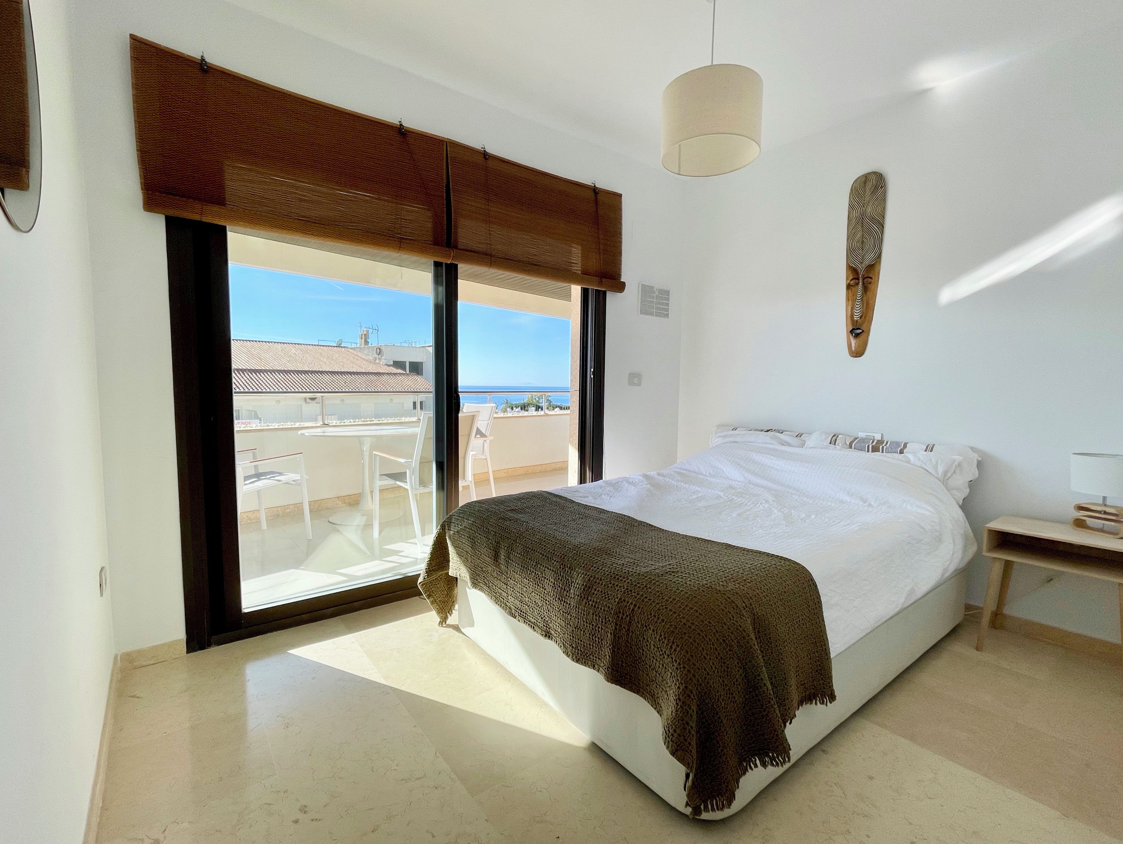 A bedroom with a bed, a nightstand, a lamp, a window with a view of the sea, and a ceiling fan.