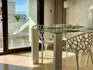 Zona pranzo moderna con tavolo in vetro e sedie bianche, con vista su un balcone e sul mare.