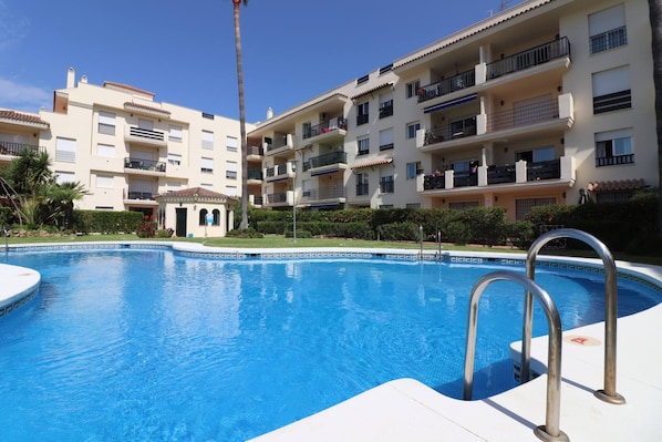 Une piscine entourée de barrières métalliques, elle-même bordée d’un immeuble doté de nombreux balcons, le tout sous un ciel d’un bleu limpide.