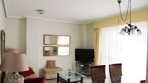 Ein Wohnzimmer mit einer Couch, einem Glastisch, einem Fernseher und einem Esstisch mit Stühlen.