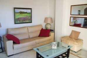 A living room with a beige sofa, red pillows, a glass coffee table, a lamp, and a framed landscape painting. - Apartment 10 Minutes Walk From Puerto Banús (Marbella)
