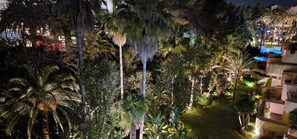 Vue nocturne d’un hôtel avec une piscine entourée de palmiers et d’une végétation luxuriante.