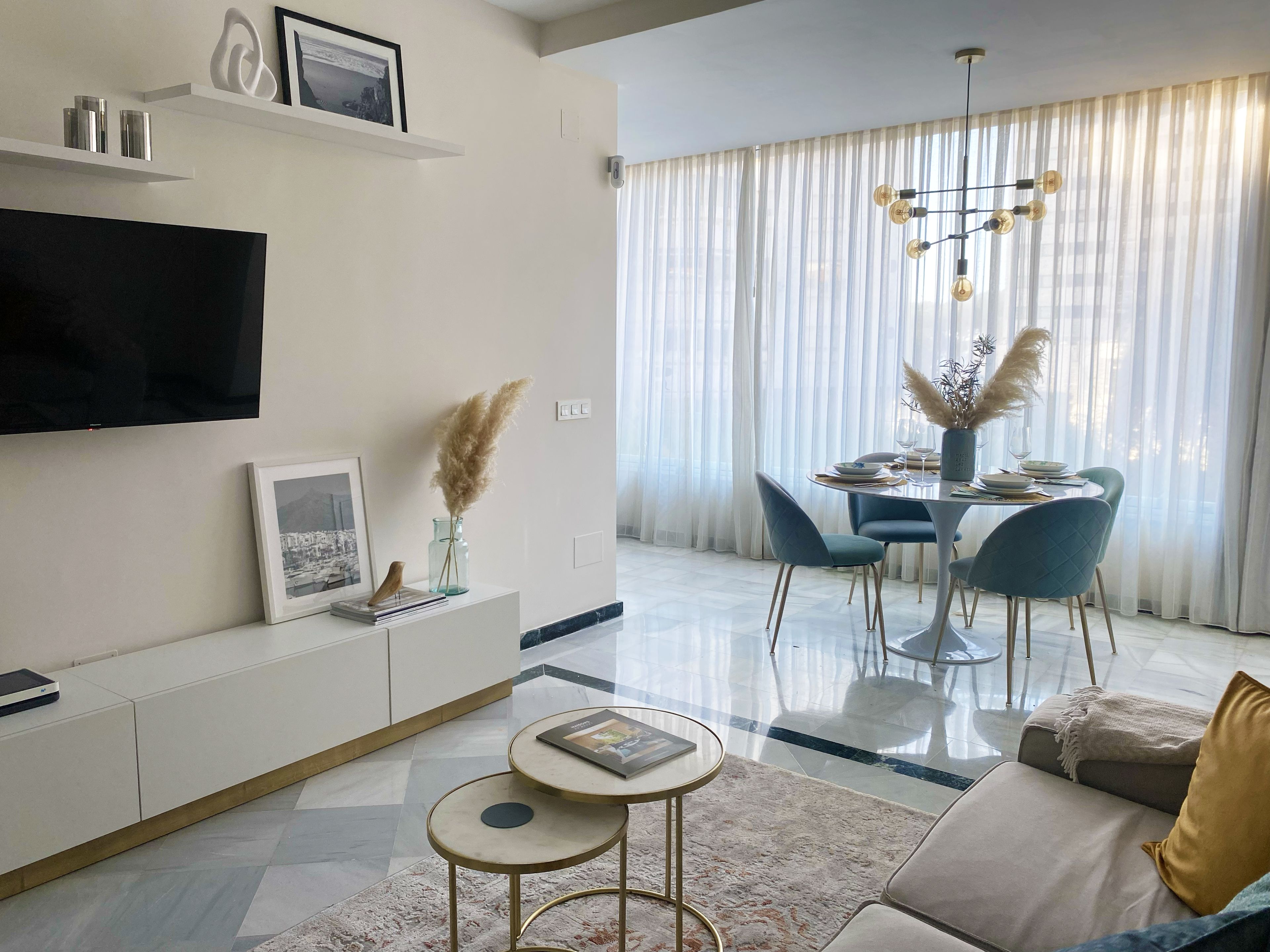 A modern living room with a flat-screen TV, a dining area with a chandelier, and a sofa with a throw blanket.