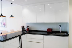 Une cuisine moderne avec des meubles blancs, un plan de travail noir et un grille-pain rouge. On y trouve deux lampes suspendues et une machine à café posées sur le plan de travail.
