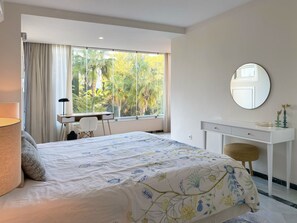 Ein Hotelzimmer mit Bett, Schreibtisch, Stuhl, einem runden Spiegel und Blick auf Palmen durch große Fenster.