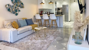 Ein modernes Wohnzimmer mit einer Couch, einem Couchtisch und Barhocker. Im Hintergrund ist eine Küche mit Kühlschrank und Mikrowelle erkennbar.