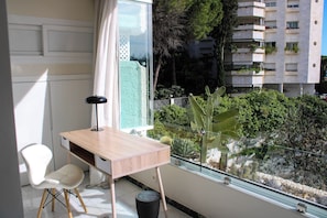 Une chambre moderne avec un bureau en bois, une chaise blanche et une grande fenêtre donnant sur un jardin et un immeuble.