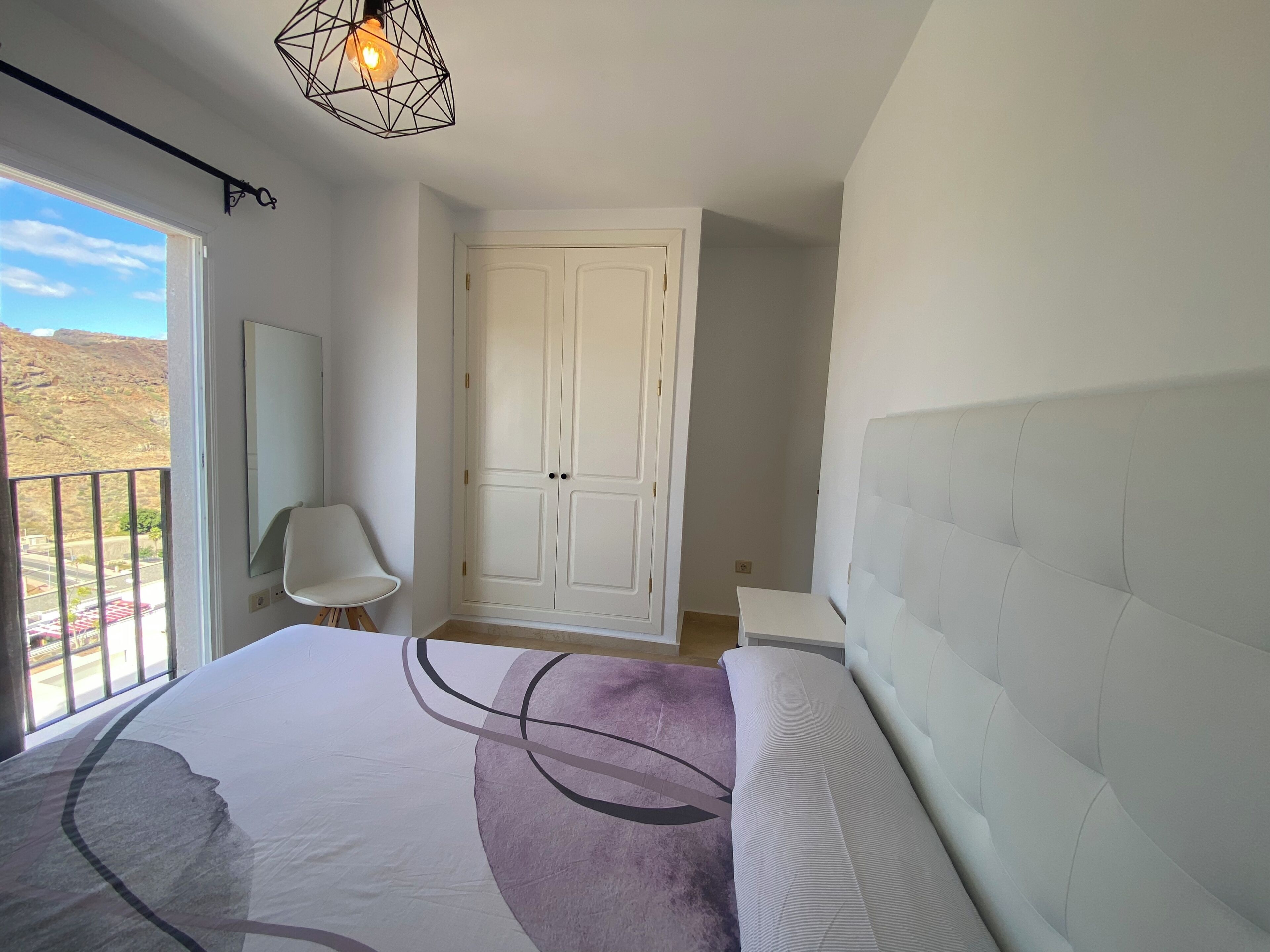 A bedroom with a large bed, a white wardrobe, a chair, and a view of the outdoors.