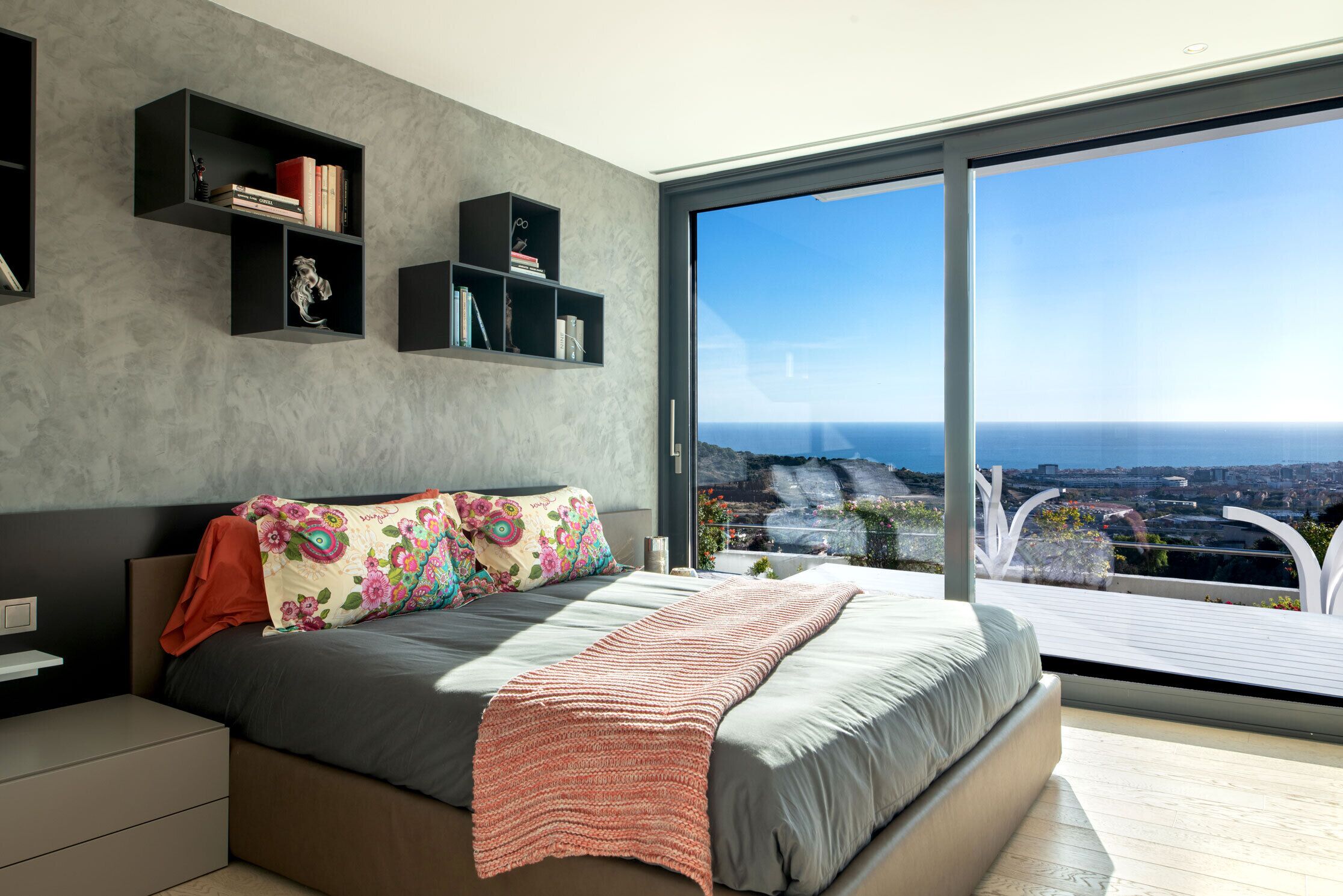 A modern bedroom with a large bed, built-in shelves, and a city view through large windows.