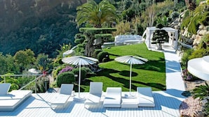 Un espace extérieur luxueux, aménagé avec des fauteuils de jardin blancs et des parasols, entouré de jardins parfaitement entretenus et offrant une vue pittoresque sur une colline.