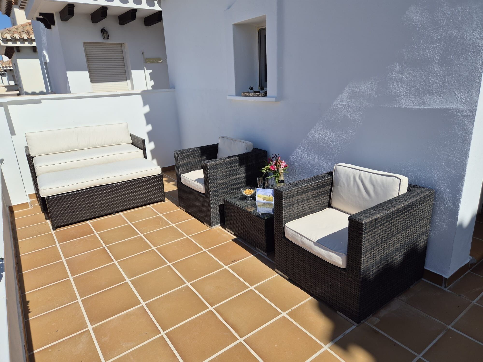 Un patio con muebles de mimbre y un sofá blanco, una pequeña mesa con un jarrón de flores, y una ventana con vistas a la terraza.