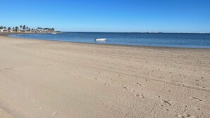 A wide sandy beach with footprints leading to the water, a small boat, and a distant shoreline with buildings and palm trees. - The Fairways - Luxurious Front Line Villa on Mar Menor Golf Resort (Torre-Pacheco)
