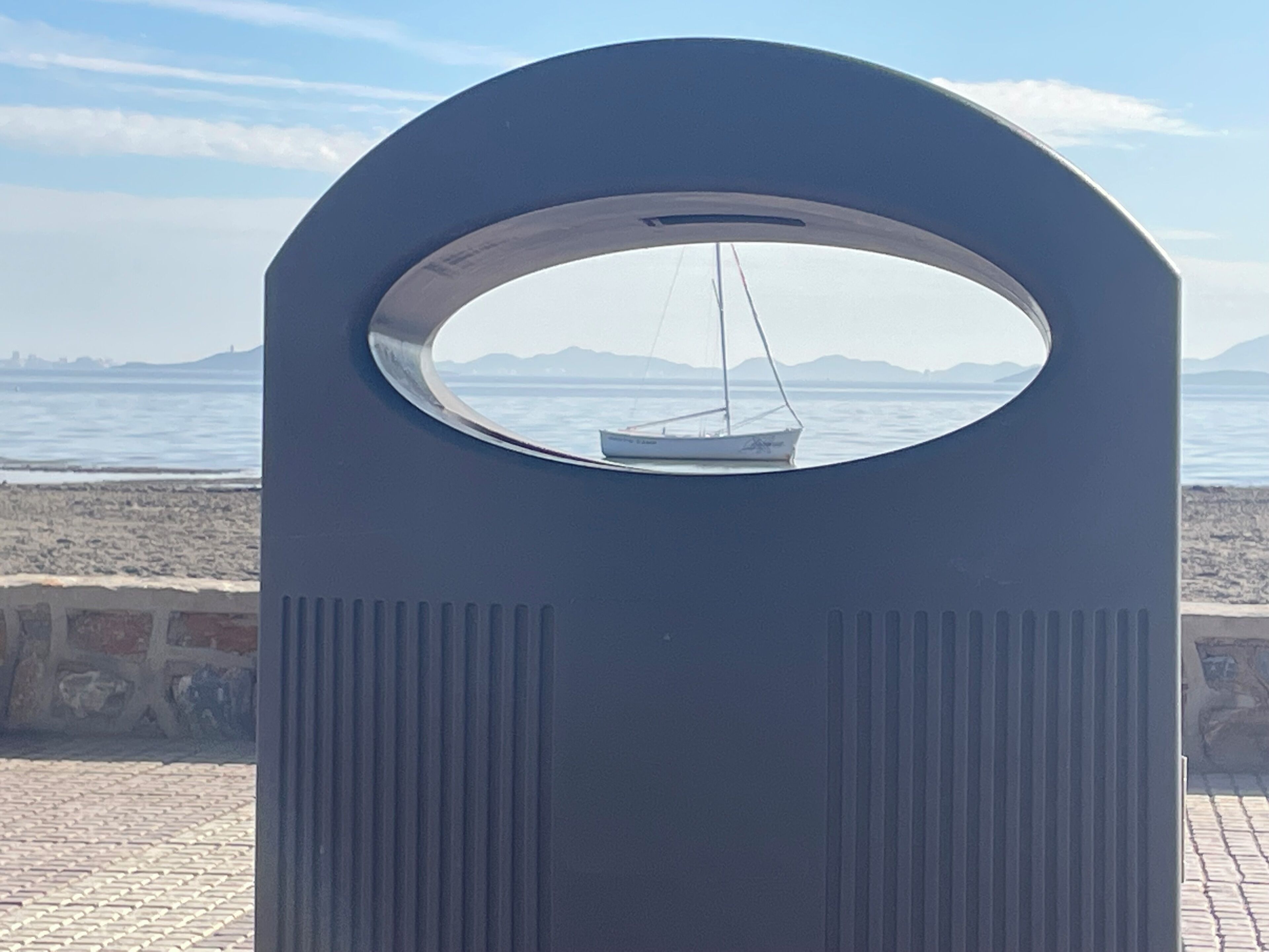 A view through a circular window of a sailboat on calm waters, with distant mountains and a clear sky.