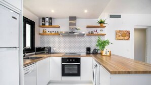 A modern kitchen with white cabinetry, a wooden countertop, a built-in oven, a dishwasher, and a coffee maker. There are shelves with jars and a plant on the wall.