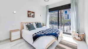 A modern bedroom with a large bed, a nightstand, a wall-mounted light, a window with a view of buildings, and a balcony with a wicker basket.