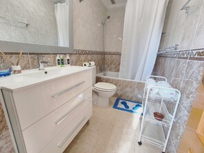 A bathroom with a white vanity, a toilet, a shower, and a small shelf with toiletries. - Sea View Bungalow With Pool (Torrevieja)