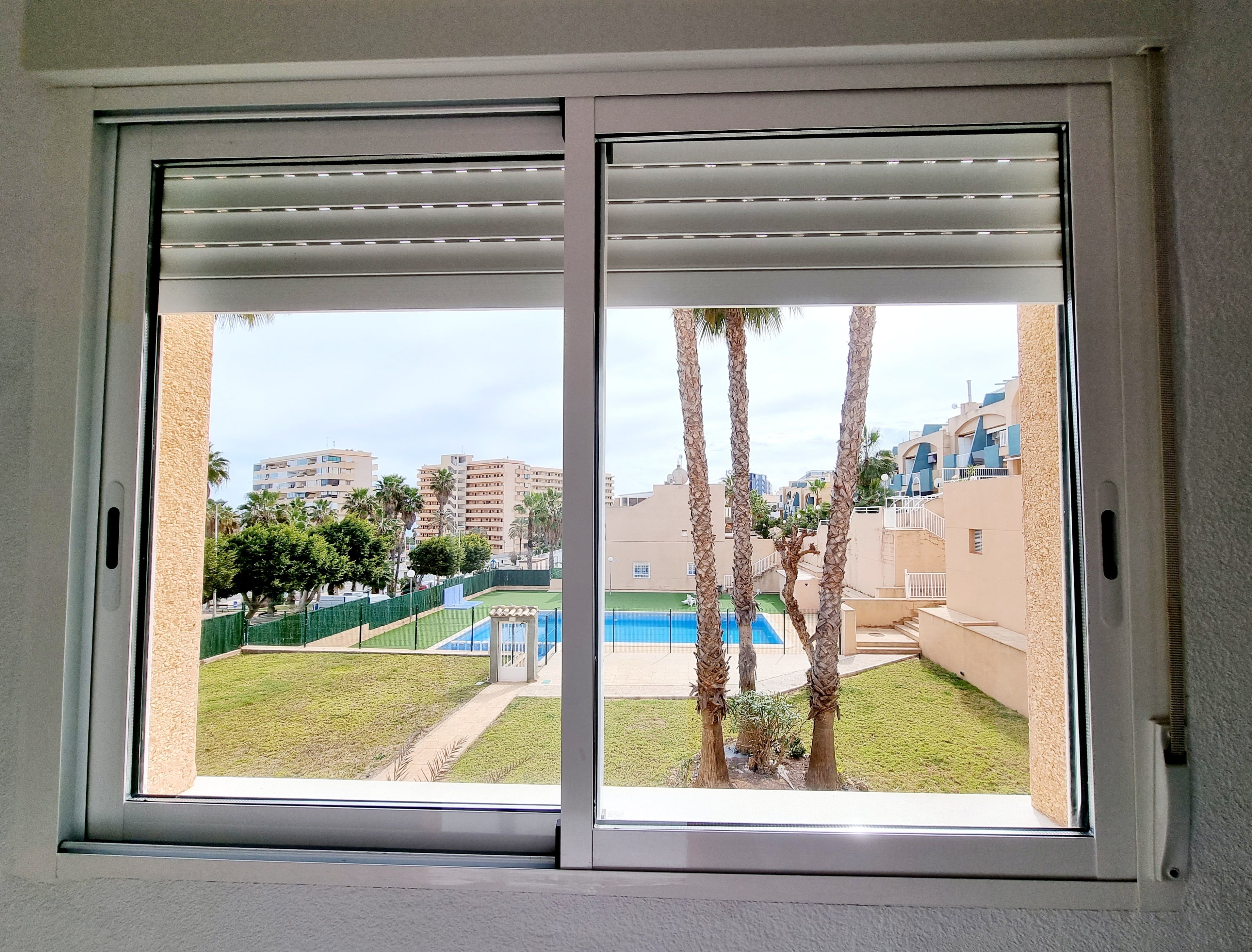 A view through a window with a sliding door, revealing a pool, palm trees, and buildings in the background.