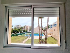 A view through a window with a sliding door, revealing a pool, palm trees, and buildings in the background. - Sea View Bungalow With Pool (Torrevieja)
