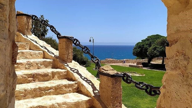 Eine Steintreppe führt zu einem Meerblick, mit einem Kettenzaun und wolkenlosem, blauem Himmel.