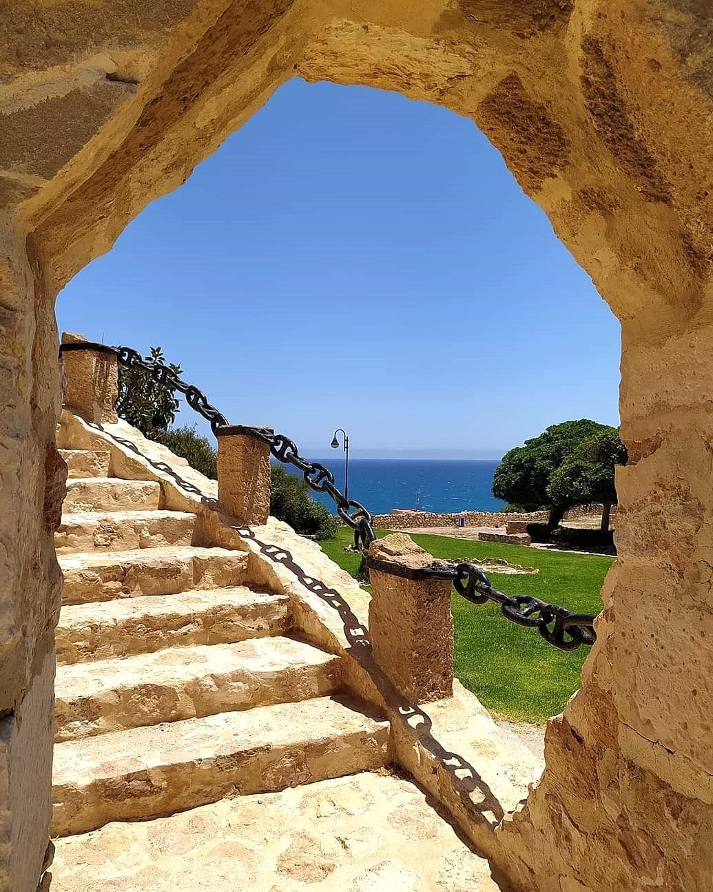 A stone staircase leading to a view of the sea, with a chain barrier and a clear blue sky.