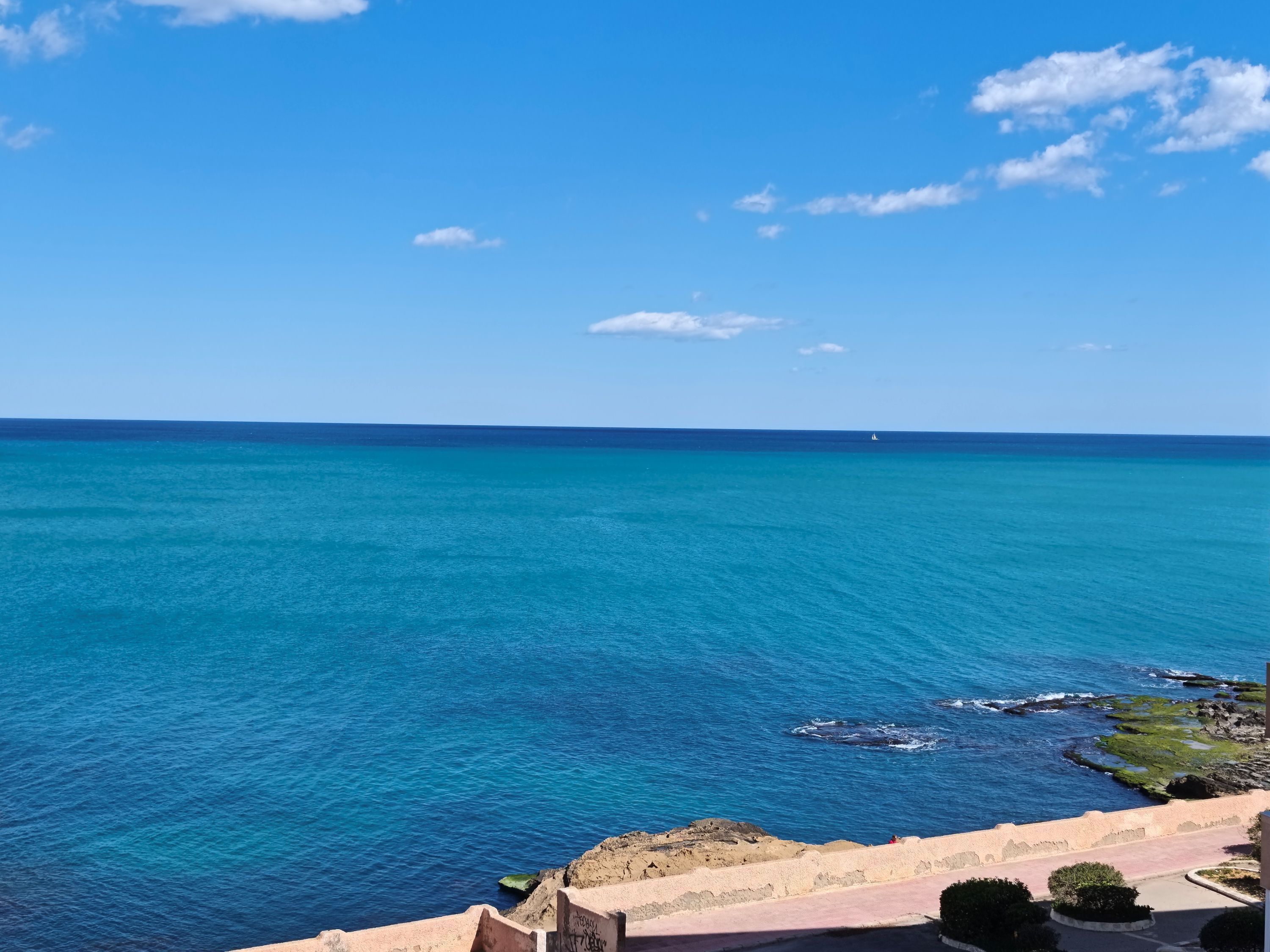 Une vue sur la côte, avec une eau d’un bleu limpide, un voilier au loin et une côte rocheuse bordée d’une promenade pavée.