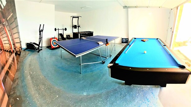 Une pièce avec une table de billard bleue, une table de ping-pong, du matériel d’exercice et une cible au sol.